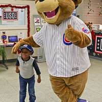 A U.S. Army Soldier's son poses with Clark, the Chicago Cubs mascot, at the  USO at USAG Bavaria in Grafenwoehr, Germany, Dec. 11, 2018. The USO  sponsored a Chicago Cubs mascot tour